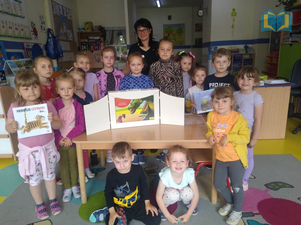 Zdjęcie: Zdjęcie grupowe dzieci w przedszkolu. Na biurku stoi biały teatrzyk Kamishibai, w której znajduje się książka pt. "Legenda o Smoku Wawelskim".